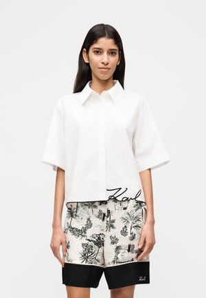 Frau mit langen dunklen Haaren, die ein weißes Kurzarm-Hemd mit Knöpfen und cremefarbene Shorts mit schwarzem tropischem Muster und schwarzem Besatz trägt.