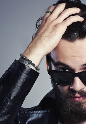 Juodas odinis švarkas, tamsiai mėlyni akiniai nuo saulės ir karoliukų apyrankė su kryžiaus detale ant riešo; šukuosena atgal ir barzda.