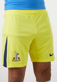 Gule atletiske shorts lavet af tekstureret stof. Har en marineblå side-stripe og broderet logo med en fugl og emblem. Nike-branding til stede.