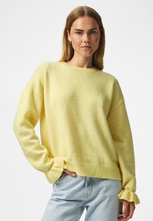 Femme aux cheveux blonds et raides portant un pull jaune doux et un jean bleu clair, debout avec une main dans la poche devant un fond blanc.