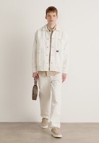 White denim jacket, beige shirt, and white pants. Wears beige sneakers, holding a gray bag. Plain background with neutral flooring.