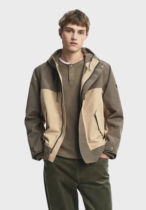 Khaki and beige color-blocked jacket with a hood, featuring a front zipper, snap buttons, and side pockets, paired with a green crewneck shirt.