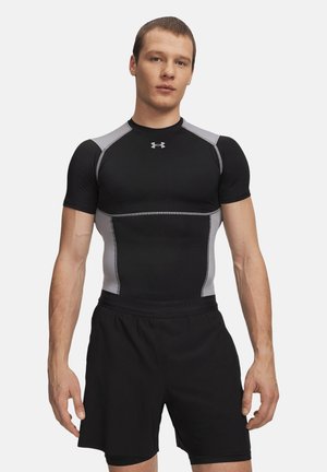 Uomo con maglietta sportiva aderente nera e grigia e pantaloncini neri, in piedi davanti a uno sfondo semplice.