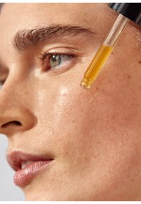 Bouteille compte-gouttes versant de l'huile jaune près d'un gros plan d'un visage lisse, de teinte claire, avec quelques taches de rousseur et un léger éclat.