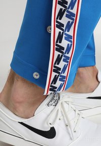 Modré sportovní kalhoty mají na boku pruh se vzorem černých a modrých prvků, červené detaily a cvoky na manžetách. Nošeny s bílými teniskami.