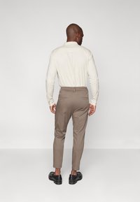 Camisa de manga larga color beige claro con tejido texturizado, combinada con pantalones marrones a medida y zapatos negros, mostrada desde atrás.