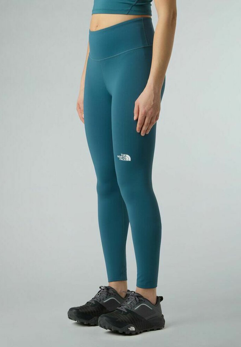 Persona che indossa leggings a vita alta color teal con il logo The North Face sulla coscia e scarpe da hiking nere, in piedi contro uno sfondo semplice.