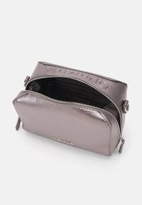 Metalická fialová kožená kabelka s kulatým tvarem, zipovým uzávěrem a reliéfním logem značky na přední straně. Uvnitř jsou přihrádky.