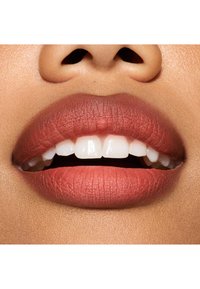 Rouge à lèvres mat couleur terre cuite appliqué sur des lèvres pleines, révélant des dents blanc nacré ; texture lisse avec des contours des lèvres définis et une légère brillance.