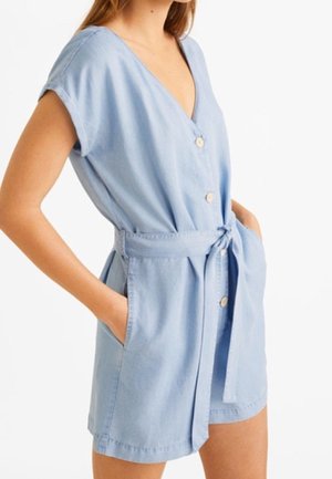 Nainen, joka pukeutunut vaaleansiniseen lyhythihaiseen napilliseen romppiin, jossa on vyötärönauha ja kädet sivutaskuissa, valkoista taustaa vasten.