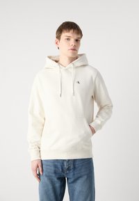Sudadera blanca hecha de una mezcla de algodón, con un bolsillo frontal, capucha con cordón y un pequeño logo negro en el pecho. Combinada con jeans de mezclilla azul.