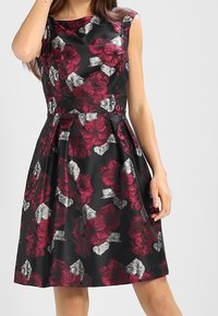 Květinové šaty s černou tkaninou, na které jsou bohaté burgundské květy a šedé listy, přiléhavý korzet a rozšířená sukně sahající až do poloviny lýtek.