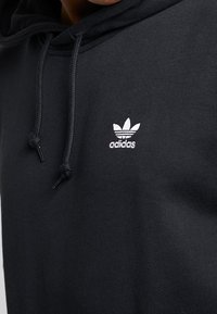 Černá mikina s kapucí na šňůrku, měkká textura a bílý vyšitý logo Adidas na levé horní části hrudi.