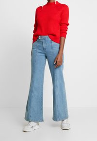 Červený žebrovaný pletený svetr ve spojení se světlými modrými rozšířenými džíny. Bílé tenisky doplňují outfit. Svetr má vysoký výstřih a texturovaný vzor.