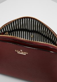 Burgunderfarbene Kosmetiktasche mit Reißverschluss, gepolstert und mit gestreiftem Stoffinnenfutter sowie goldenen Beschlägen. Verfügt über das Markenlogo auf der Vorderseite und eine glatte Außenfläche.