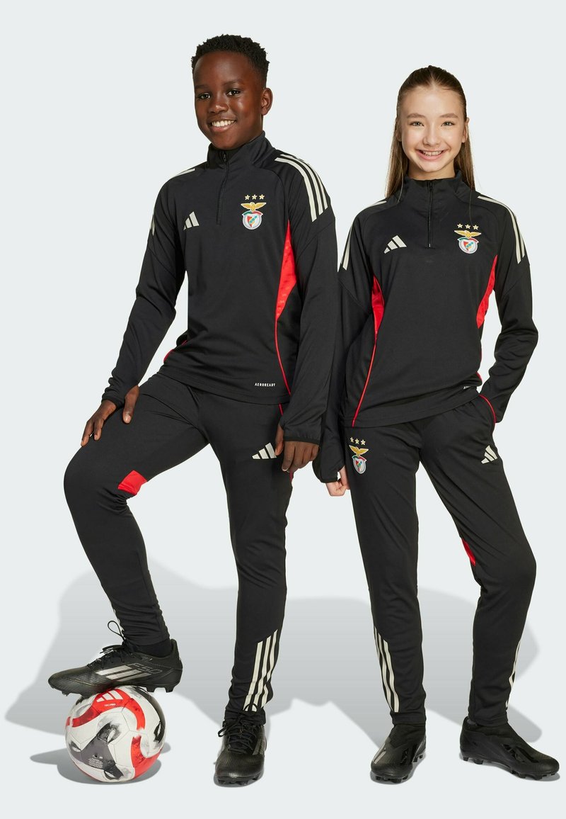 Tute sportivi neri con accenti rossi, dotati di colli con zip e del logo del Benfica. Il modello posa con un pallone da calcio e scarpini.