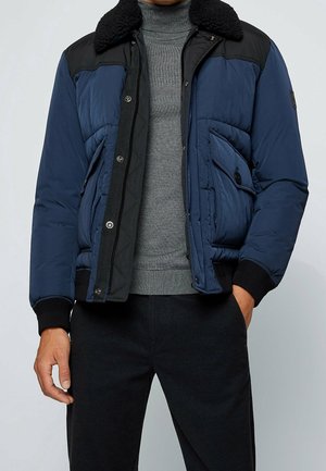 Homme portant une veste matelassée bleue et noire avec un col en peau lainée sur un pull gris à col roulé et un pantalon noir.