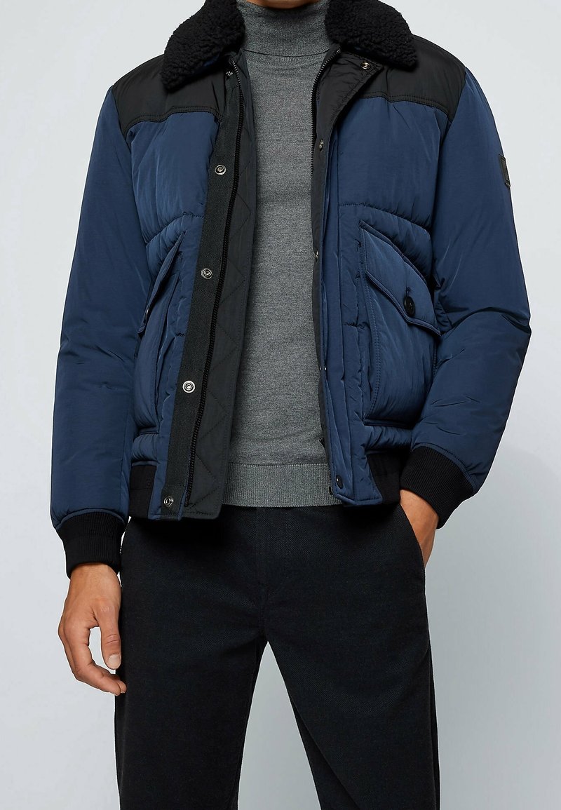 Homme portant une veste matelassée bleue et noire avec un col en peau lainée sur un pull gris à col roulé et un pantalon noir.