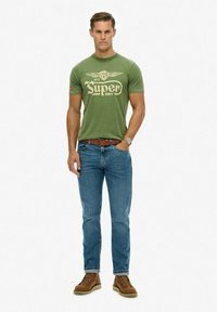 Camiseta de algodón verde con el logo "Super Dry", combinada con jeans azules y botas marrones con cordones. El modelo está de pie con las manos en los bolsillos.