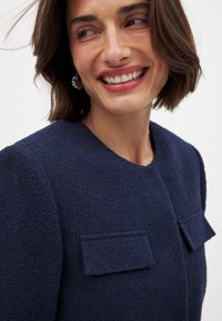 Jūros mėlynos tekstūruotos striukės su apvaliu iškirpimu, dviem kišenėmis priekyje ir trumpomis rankovėmis. Matomas lygaus audinio detalės.