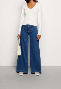 Weißes, geripptes Langarmoberteil mit V-Ausschnitt und Knopfdetails in der Mitte, kombiniert mit weit geschnittenen blauen Jeans und einer weißen Handtasche.