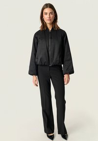Černá bomber bunda s hladkou texturou, zapínáním na zip a širokými rukávy, spárovaná s fitted černými kalhotami a jehlovými podpatky.