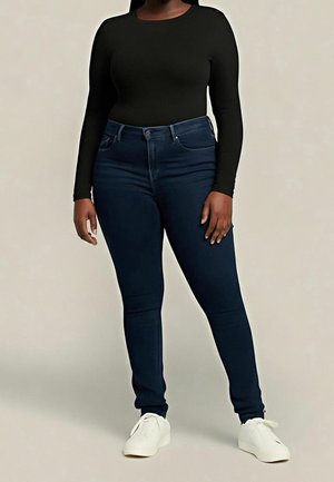 Osoba oblečená v černém tričku s dlouhými rukávy, tmavě modrých úzkých džínách a bílých teniskách stojí před jednoduchým pozadím.