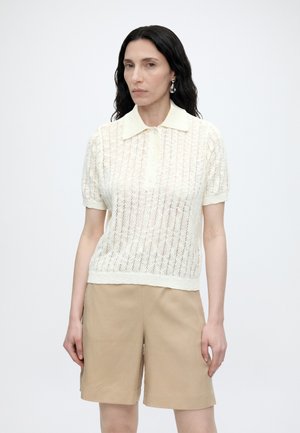 Femme aux longs cheveux noirs portant un polo en maille blanc à manches courtes et un short beige mi-cuisses sur un fond uni.