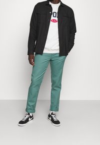 Chemise noire boutonnée, sweat-shirt gris avec logo, pantalon bleu sarcelle, baskets noires et blanches. Tissu lisse, coupe décontractée et revers de pantalon roulés.