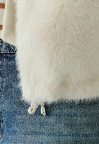 Witte nepbontstof met een zachte textuur, voorzien van een trekkoord aan de onderkant met zilverkleurige hardware, tegen een achtergrond van blauwe denim.
