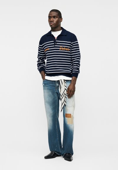 Jeune homme portant un polo à manches longues bleu marine rayé, un jean délavé avec des patchs et une écharpe rayée pendante, ainsi que des chaussures noires pointues, debout devant un fond blanc.