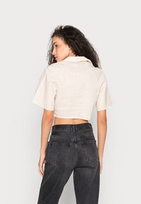 Camisa de linho beige, cortada, com gola e mangas curtas, combinada com jeans justos de ganga pretos de cintura alta, com bolsos nas costas.