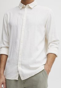 Chemise blanche en lin à boutons avec un col décontracté, manches longues retroussées jusqu'aux coudes, et texture subtile. Associée à un pantalon gris clair.