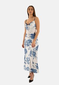 Robe midi florale avec des motifs bleus sur fond blanc, dotée de fines bretelles et d'un décolleté en dentelle à bords festonnés, associée à des talons noirs.