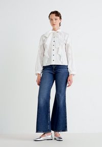 Mujer vestida con una blusa blanca con volantes y botones negros, jeans de pierna ancha de color azul oscuro y bailarinas plateadas, de pie contra un fondo blanco liso.