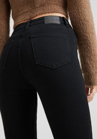 Vaqueros ajustados negros de mezclilla, con una cintura de altura media, bolsillos traseros y un suéter marrón texturizado sobre la cintura.