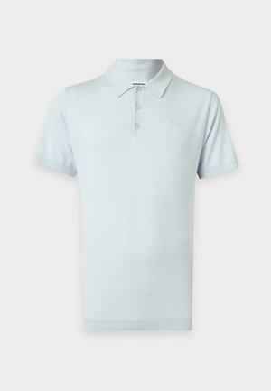 Hombre que lleva una camiseta polo azul claro de manga corta, vaqueros azul claro y zapatillas blancas, de pie sobre un fondo blanco liso.