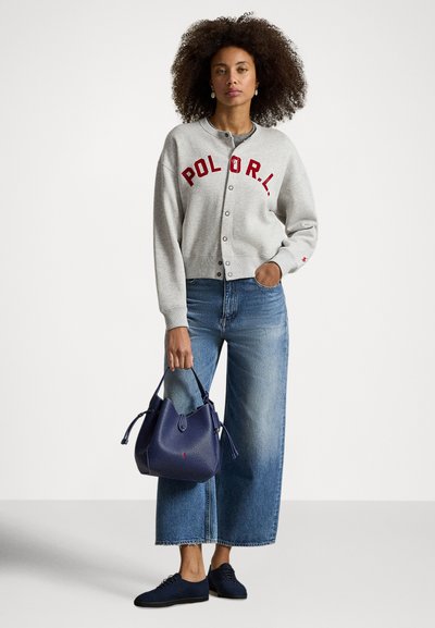 Mulher com cabelo encaracolado, vestindo um casaco cinzento com botões e letras vermelhas, jeans azuis de perna larga, sapatos pretos, segurando uma mala azul-marinho.