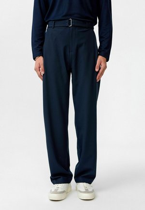 Marineblauwe broek met een rechte pasvorm, voorzien van een platte voorkant, riemlussen en een gladde, gestructureerde stof. Draagt met witte sneakers.