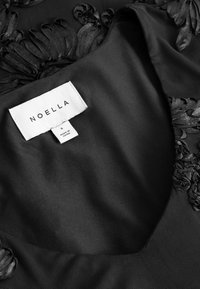Zwarte stof met geborduurde bloemenpatronen en een wit kledinglabel met de tekst "NOELLA," maat S, gemaakt in China.