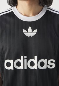 Černé sportovní tričko s vertikálními pruhy, bílým límcem a třemi bílými pruhy na ramenou. Má velké bílé logo "adidas" a ikonické logo.