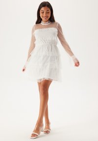 Vit lager-på-lager klänning i prickig tyl, med långa genomskinliga ärmar och en volangkrage. Bärs med klara klacksandaler.