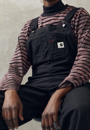 Osoba ubrana w czarne ogrodniczki Carhartt na fioletowo-czarnej, paskowanej koszulce z długim rękawem, siedząca na tle szarego, teksturowanego tła.