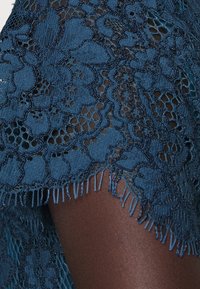 Spetsöverdrag i djup teal med intrikata blommönster, som har fransade kanter för textur och detaljer. Halvgenomskinlig look.