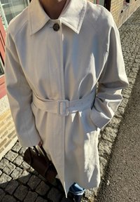 Jasbeżowy trencz o zróżnicowanej strukturze, z paskiem w talii, zapięciem na guziki i dwiema bocznymi kieszeniami. Noszony z niebieskimi dżinsami, trzymany w brązowej torbie.