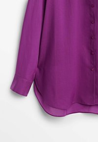 Chemise violette à manches longues avec ourlet arrondi et fermeture boutonnée sur le devant, montrant la manche gauche et la partie inférieure du torse.