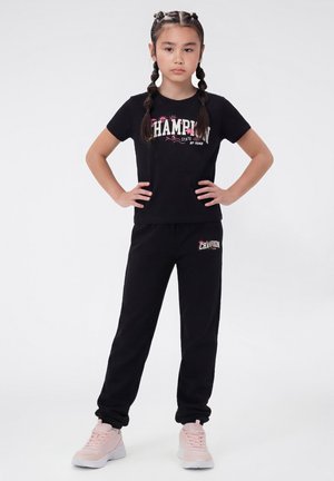 Junges Mädchen steht mit den Händen in den Hüften, trägt ein schwarzes "Champion"-T-Shirt und Hose sowie hellrosa Turnschuhe vor weißem Hintergrund.