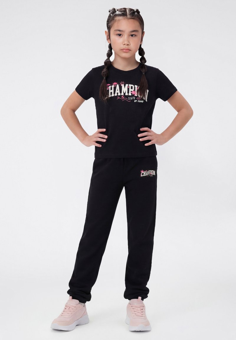 Jeune fille debout, mains sur les hanches, portant un t-shirt noir "Champion" et un pantalon, ainsi que des baskets rose clair sur un fond blanc.