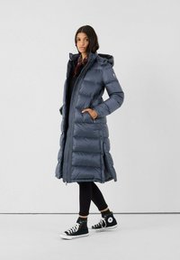 Giacca a vento imbottita di un grigio scuro con zip frontale, cappuccio e tasche laterali; presenta un design lungo e aderente con texture trapuntata e dettagli in bottone.