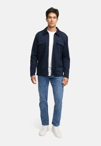 Mørkeblå jakke med to brystlommer, krave og lynlås foran, parret med lyseblå jeans og hvide sneakers.